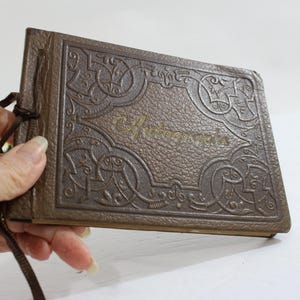 May include: A brown, vintage-style photo album with embossed decorative patterns and the word "Photographs" in gold lettering. The album is bound with a brown ribbon. The album is held by a person's hand.