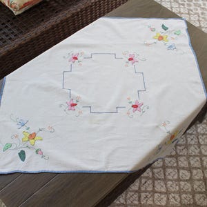 May include: A white square tablecloth featuring a central blue embroidered geometric frame, surrounded by four pink floral motifs. The corners are decorated with yellow and green floral patterns, and the edges are finished with a blue trim.