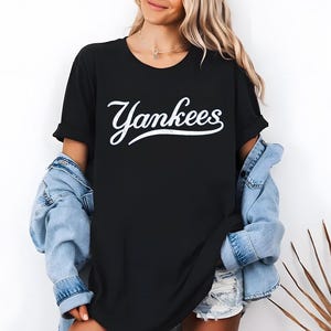 May include: Black t-shirt with the word "Yankees" in white script. The shirt has short sleeves and a crew neck. A light blue denim jacket is worn over the shirt.