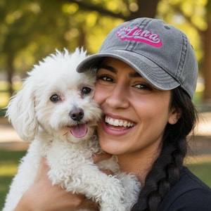 Könnte beinhalten: Graue Baseballkappe mit dem Namen "Luna" in rosa Schrift. Eine Person hält einen kleinen, flauschigen weißen Hund. Der Hintergrund ist ein Park mit grünen Bäumen und Gras.