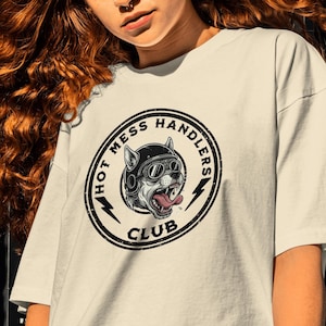 May include: Cream-coloured t-shirt with a circular graphic that reads "HOT MESS HANDLERS CLUB." The graphic depicts a dog wearing a helmet and sunglasses, with its tongue out. The shirt has short sleeves and a relaxed fit.