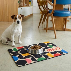 Könnte beinhalten: Ein Hund sitzt neben einer bunten Futtermatte für Haustiere mit einem geometrischen Muster aus schwarzen, blauen, gelben, grünen und rosa Formen. Eine Edelstahlschüssel mit Hundefutter steht auf der Matte. Auf der Matte steht der Name "MILO" in Rot.