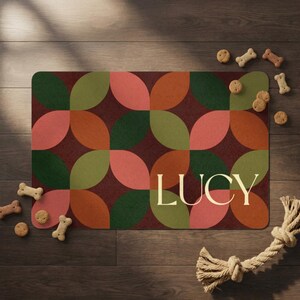 May include: A rectangular doormat with a geometric pattern of overlapping shapes in shades of green, orange, and pink on a burgundy background. The name "LUCY" is displayed in a cream-colored font. Dog treats and a rope toy are scattered around the mat.
