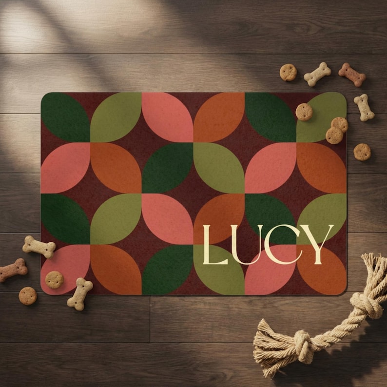 May include: A rectangular doormat with a geometric pattern of overlapping shapes in shades of green, orange, and pink on a burgundy background. The name "LUCY" is displayed in a cream-colored font. Dog treats and a rope toy are scattered around the mat.