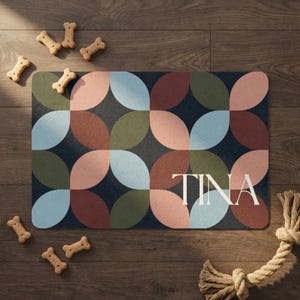 May include: A rectangular pet food mat with a geometric pattern in shades of blue, green, pink, and brown. The name "TINA" is printed in white. Several bone-shaped dog biscuits and a rope toy are also visible.