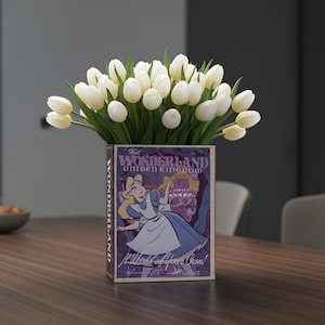 May include: A floral arrangement featuring white tulips in a rectangular vase. The vase is decorated with a vintage-style illustration of Alice in Wonderland, with text that reads "Visit Wonderland United Kingdom" and "A World of Your Own!"
