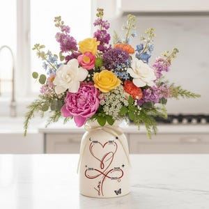 May include: A vibrant floral arrangement featuring a variety of colorful blooms, including yellow roses, pink peonies, and white gardenias, in a cream-colored vase. The vase has a heart design with dates and the word "Love".