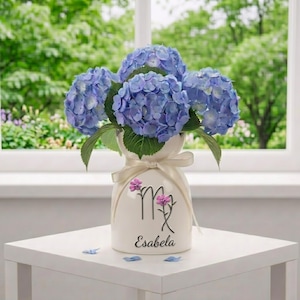 May include: A white ceramic vase with a cream-colored ribbon holds a bouquet of blue hydrangeas. The vase has a floral design and the name "Esabela" written on it. The vase is on a white table in front of a window.