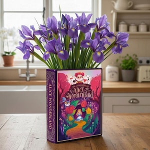 May include: A purple ceramic vase shaped like a book, featuring the cover art of "The Adventures of Alice in Wonderland," holds a bouquet of purple irises. The vase is on a wooden surface, with a kitchen in the background.