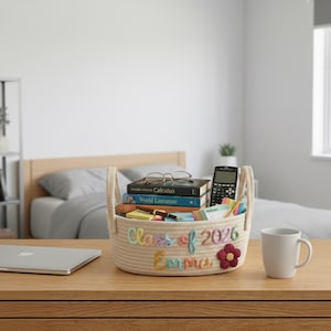 May include: A woven rope basket with handles, filled with books, pens, a calculator, and notepads. The basket is personalized with the text "Class of 2026 Emma" and a flower. A white mug and laptop are on the wooden surface.