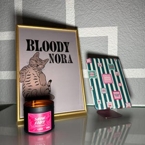 Peut inclure: Une affiche encadrée avec le texte "BLOODY NORA" et une illustration de chat, une bougie noire avec une étiquette rose "SNOW FAIRY", et un carnet à rayures avec des motifs de timbres.