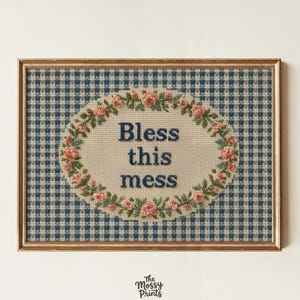 May include: A rectangular framed cross-stitch embroidery featuring a blue and white gingham background. An oval center contains the text Bless this mess in dark blue letters, surrounded by a decorative wreath of small pink roses and green leaves.