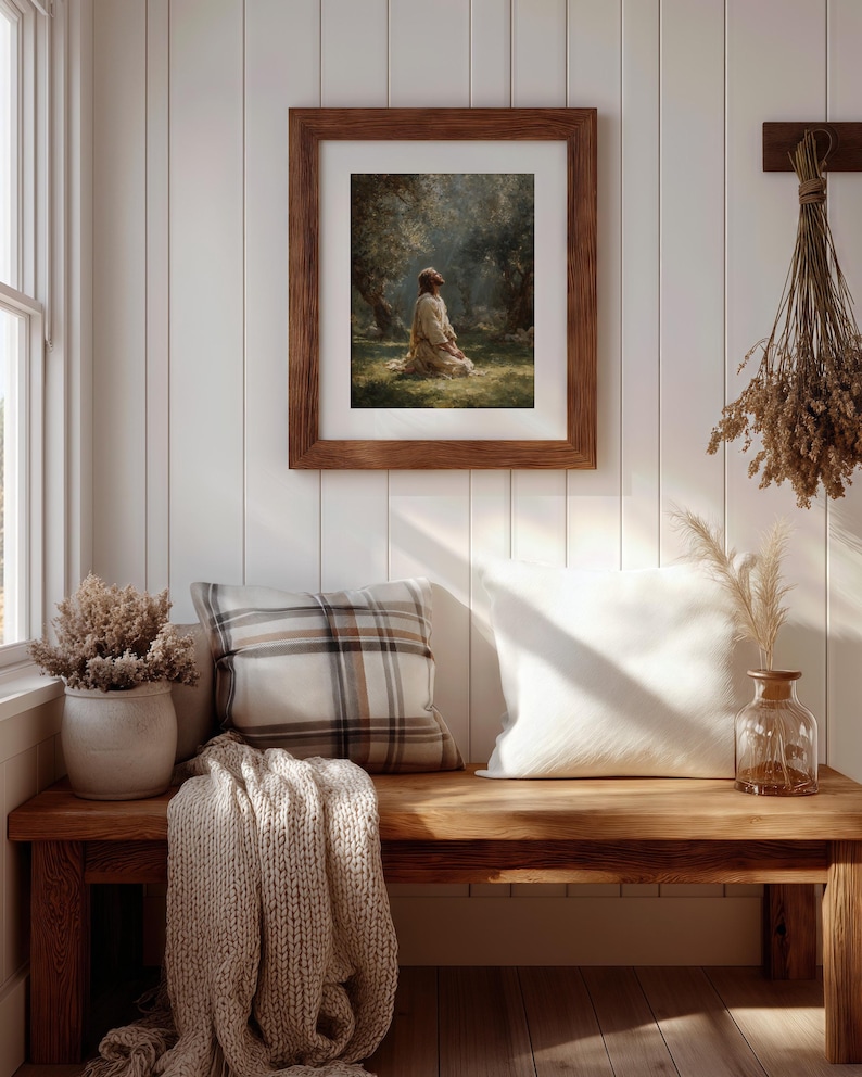 Peut inclure: Une &oelig;uvre d'art encadr&eacute;e repr&eacute;sente une personne agenouill&eacute;e dans une for&ecirc;t ensoleill&eacute;e. L'&oelig;uvre est dans un cadre en bois marron, accroch&eacute;e &agrave; un mur &agrave; panneaux blancs. Un banc en bois contient des coussins, une couverture tricot&eacute;e et des vases d&eacute;coratifs avec des plantes s&eacute;ch&eacute;es.