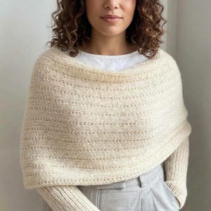 May include: A cream-coloured knitted poncho featuring a textured horizontal stitch pattern. The garment has a wide, rounded neckline and long sleeves that extend from the main body, crafted from a soft, thick yarn with a subtle heathered appearance.