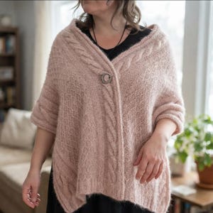 May include: A dusty pink knitted poncho featuring a textured honeycomb pattern and vertical cable knit borders along the front opening. The garment is secured at the neckline with a circular metal clasp and has short, wide sleeves.