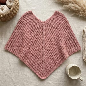 May include: A dusty rose knitted poncho featuring a V-shaped neckline and a central vertical ribbed seam detail. The garment is crafted with a textured garter stitch pattern, creating a soft, uniform appearance across the entire triangular silhouette.