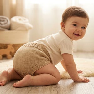 May include: A beige knitted diaper cover featuring a ribbed waistband and leg openings, paired with a short-sleeved cream-colored ribbed shirt. The garment set is worn by a small child who is positioned on all fours on a light-colored surface.