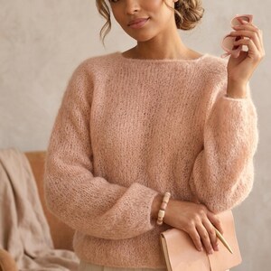 May include: A soft, fuzzy, light pink long-sleeved sweater with a wide boat neckline. The knit fabric has a loose, airy texture with visible fibers. The sweater features raglan sleeves and a relaxed fit, providing a comfortable and casual appearance.