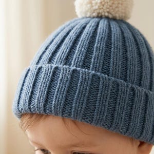 Può includere: Un berretto in maglia a coste blu con un bordo spesso ripiegato e un grande pompon rotondo color crema sulla parte superiore. Il berretto presenta una trama a coste verticali su tutto il corpo principale e sul bordo, creando un motivo.