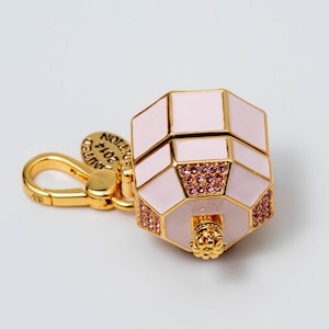 May include: A gold-toned metal charm shaped like a small, octagonal perfume bottle with pale pink enamel panels. The piece features small pink crystals embedded on the sides and a gold lobster clasp with a round tag reading LIMITED EDITION 2014.