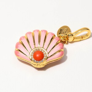 May include: A gold-tone metal charm shaped like a seashell with light pink enamel detailing. The center features a round orange stone surrounded by a ring of small clear crystals. A gold-tone lobster clasp is attached to the top of the shell.
