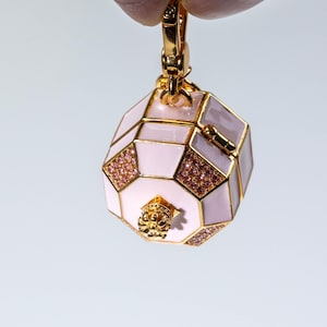 May include: A gold-toned charm in the shape of an octagonal box with light pink enamel panels, gold metal trim, and small pink crystal accents. The charm features a small gold crown-like detail on the bottom and a gold lobster clasp at the top.