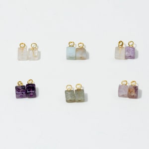 Puede incluir: Seis pares de pequeños dijes de piedras preciosas rectangulares, cada uno con una anilla de metal dorado en la parte superior. Las piedras varían en color, incluyendo tonos transparentes, azul claro, morado, morado oscuro, verde y rosa.