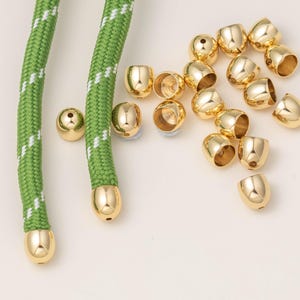 May include: A collection of small, polished gold-tone metal cord end caps, with two caps attached to the ends of a green braided cord featuring white diagonal stitching. The remaining loose caps are scattered to the right of the cord ends.