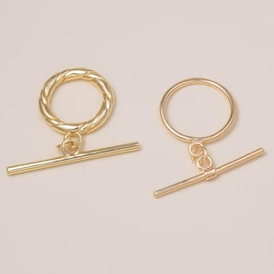 May include: Two gold-toned toggle clasps for jewelry making. One clasp features a twisted circular ring and a straight bar, while the other consists of a smooth circular ring and a straight bar with a small coiled detail at the center.