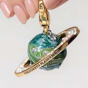 May include: A gold-coloured charm featuring a planet design. The planet is green and blue with a gold ring encrusted with small, clear stones. The charm has the text "WELCOMTO PLANETJUICY" in gold lettering.
