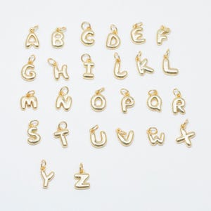 Op de afbeelding: Een verzameling van zesentwintig individuele letterbedels van goudkleurig metaal, elk met een afgeronde, bolle letterstijl en een kleine ronde ring aan de bovenkant voor bevestiging. De set bevat alle letters van het alfabet van A tot Z.