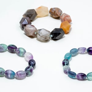 May include: Three beaded bracelets are displayed. One bracelet features large, faceted, multi-colored earth-toned stones. Two matching bracelets consist of smooth, oval-shaped beads in varying shades of translucent purple, teal, and green.
