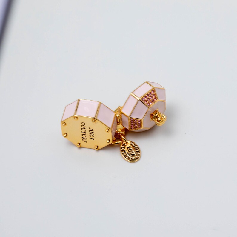 May include: A gold-tone metal charm featuring two octagonal, light pink enamel components. One side is engraved with the text JUICY COUTURE, and a small circular tag attached to the base is engraved with the text LIMITED EDITION 2014.
