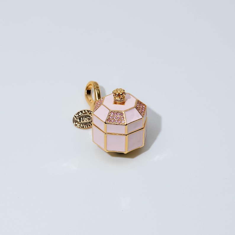 May include: A gold-toned charm shaped like an octagonal box with light pink enamel panels. The lid features a small gold crown finial and pink crystal accents. A gold lobster clasp is attached, along with a round tag engraved with 2014 EDITION.