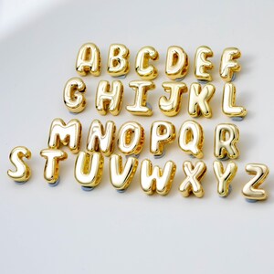 May include: A collection of twenty-six individual gold-coloured metal alphabet letter charms, arranged in rows from A to Z. Each letter features a smooth, rounded, balloon-like design with a reflective surface, designed as small decorative beads.
