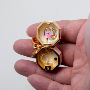 May include: A gold-toned, octagonal hinged trinket box keychain featuring a miniature pink ballerina figurine inside the top lid, a small mirror, and a bottom compartment containing a tiny pearl-like beaded bracelet and a small blue stone ring.