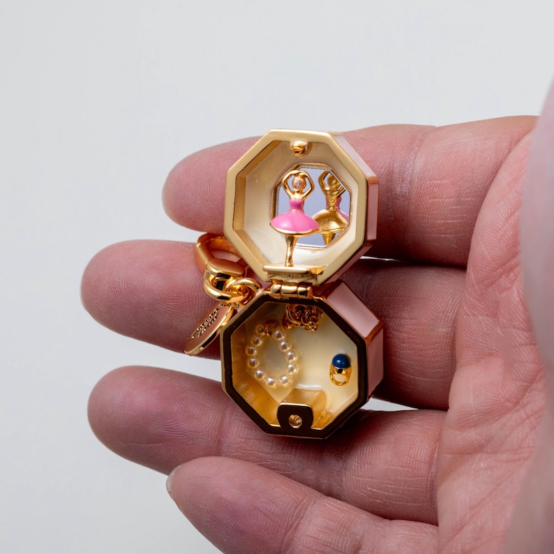 May include: A gold-toned, octagonal hinged trinket box keychain featuring a miniature pink ballerina figurine inside the top lid, a small mirror, and a bottom compartment containing a tiny pearl-like beaded bracelet and a small blue stone ring.