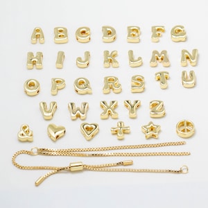 Peut inclure: Une collection de breloques en métal doré représentant les lettres de l'alphabet de A à Z, ainsi qu'une esperluette, un cœur, une croix, une étoile et un signe de paix. Sous les breloques se trouve un bracelet en chaîne boîte dorée réglable.