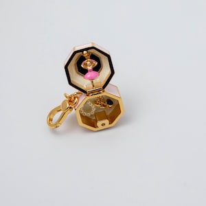 May include: A gold-toned metal charm shaped like an open hexagon box with a pale pink exterior. Inside the top section is a small pink-clad dancer figure. The bottom section contains a tiny white beaded ring and a small blue-accented gold charm.