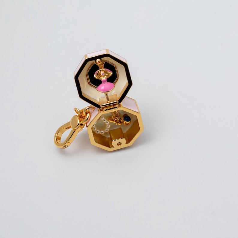 May include: A gold-toned metal charm shaped like an open hexagon box with a pale pink exterior. Inside the top section is a small pink-clad dancer figure. The bottom section contains a tiny white beaded ring and a small blue-accented gold charm.