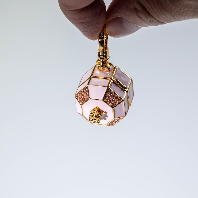 May include: A gold-toned charm in the shape of an octagonal box with light pink enamel panels, gold metal trim, and small pink crystal accents. The charm features a small gold crown-like detail on the bottom and a gold lobster clasp at the top.
