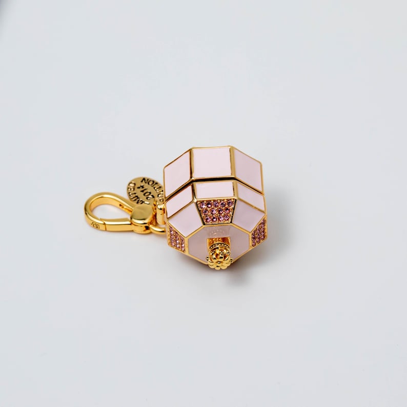 May include: A gold-toned metal charm shaped like a small, octagonal perfume bottle with pale pink enamel panels. The piece features small pink crystals embedded on the sides and a gold lobster clasp with a round tag reading LIMITED EDITION 2014.
