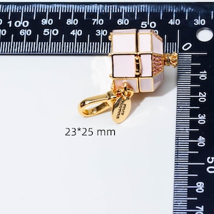 May include: A gold-tone metal charm shaped like a small box with light pink enamel panels, a crown-shaped top, and pink crystals. It features a lobster clasp and a small oval tag engraved with Juicy Couture. The charm measures 23 by 25 mm.