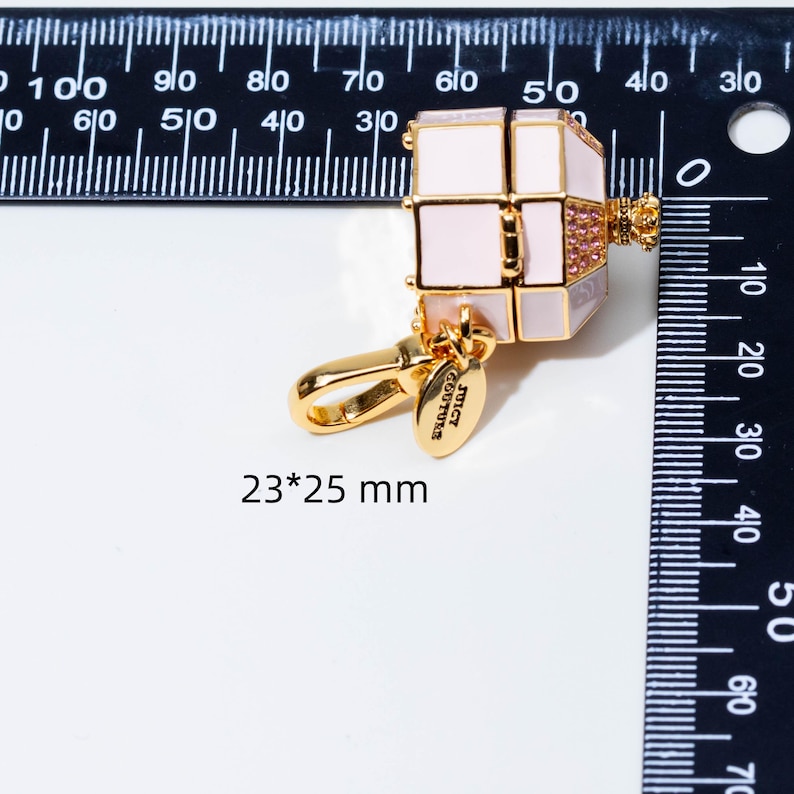 May include: A gold-tone metal charm shaped like a small box with light pink enamel panels, a crown-shaped top, and pink crystals. It features a lobster clasp and a small oval tag engraved with Juicy Couture. The charm measures 23 by 25 mm.