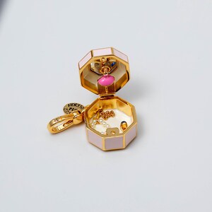 May include: A gold-tone metal charm shaped like an open octagonal box with pale pink enamel sides. The interior features a small pink figure, a delicate gold chain with white beads, and a tiny blue accent. A gold lobster clasp is attached to the side.