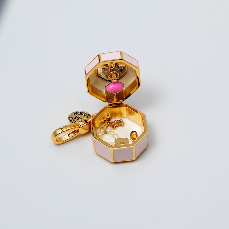May include: A gold-tone metal charm shaped like an open octagonal box with pale pink enamel sides. The interior features a small pink figure, a delicate gold chain with white beads, and a tiny blue accent. A gold lobster clasp is attached to the side.
