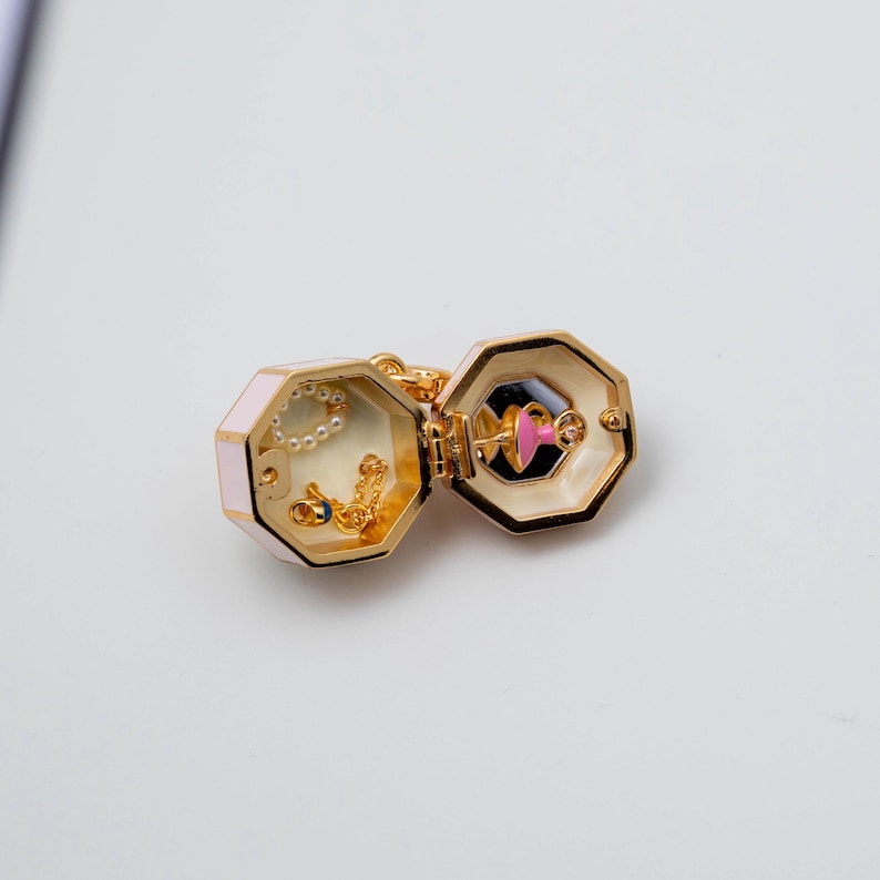 May include: A gold-toned, hinged, octagonal locket charm with a pale pink exterior. The interior features a small, pink-skirted dancer figure on one side and a delicate gold chain with a white pearl-like bead inside the other compartment.