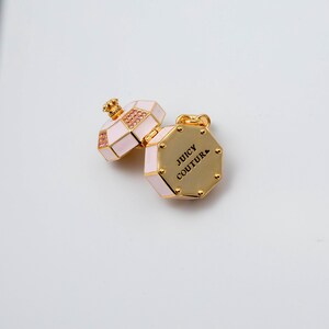 May include: A gold-tone metal charm shaped like an octagonal perfume bottle with light pink enamel panels and small pink crystals. The piece features a hinged lid and the text JUICY COUTURE engraved in black on the flat gold-tone base surface.