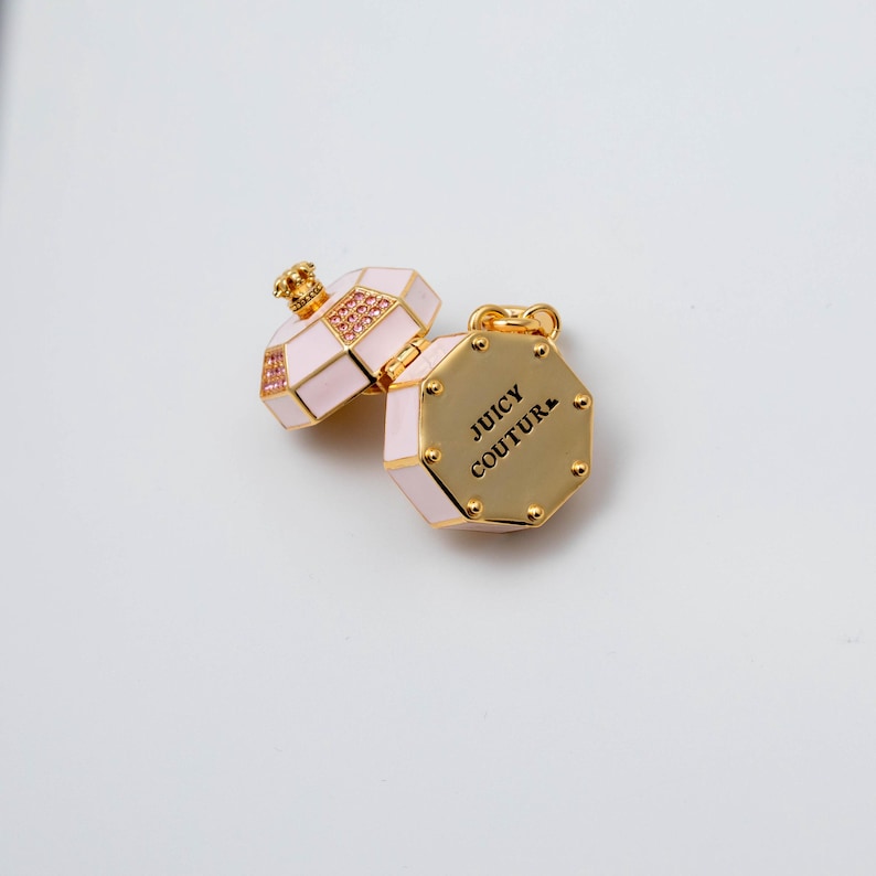 May include: A gold-tone metal charm shaped like an octagonal perfume bottle with light pink enamel panels and small pink crystals. The piece features a hinged lid and the text JUICY COUTURE engraved in black on the flat gold-tone base surface.