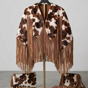 May include: A cow print jacket with long brown fringe, a matching rectangular clutch, a small shoulder bag with fringe, loafers with cow print and brown fringe, a small notebook, and two brown leather cases on a gold display stand.